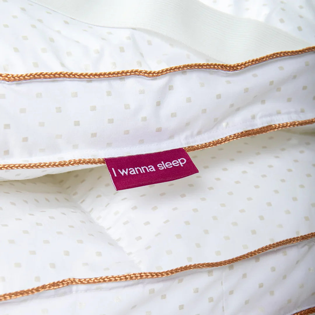 Imagem do Pillow Top Magnum aumentada destacando os detalhes de seu material e design, le possui a cor branca com pequenos quadradinhos dourados sutis, e um tecido premium com margens douradas nas laterais trazendo mais sofisticação, mais ao centro da imagem podemos ver a etiqueta ta i wanna sleep de forma minimalista